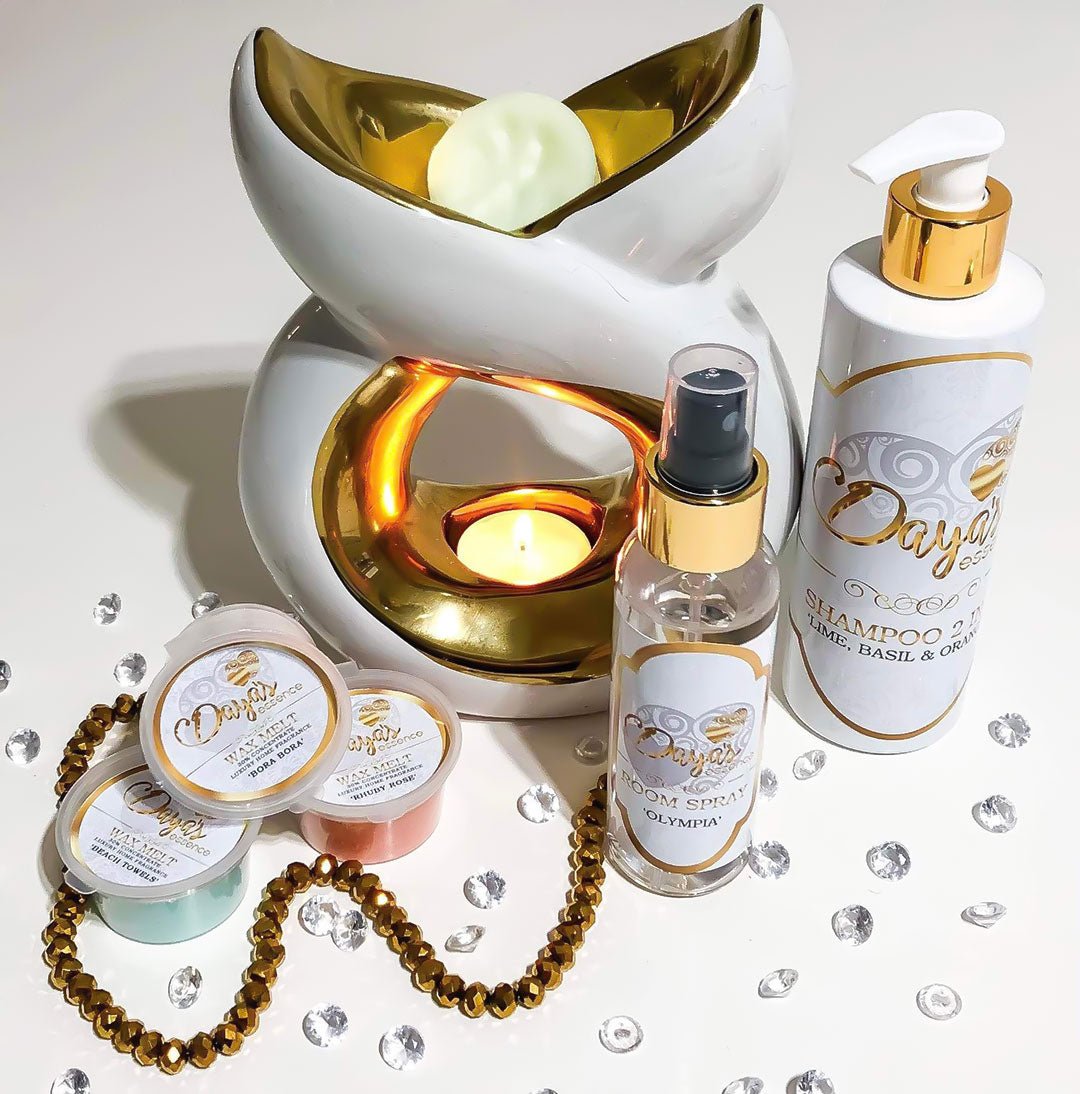 A white and gold wax melt warmer with a tealight glows softly, surrounded by Daya's Essence wax melts, a room spray, and shampoo bottle with dayas essence logo, this blog explains the difference between handmade and mass produced, which is better.