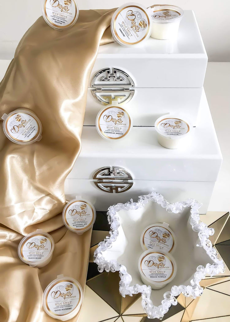 Daya's Essence wax melts in various scents, including Dove Supreme and Beach Towels, displayed on a white and gold fabric background with white decorative boxes.