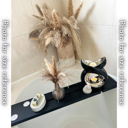 A black bath tray across a bathtub holds a bar of soap on a white dish, a vase with dried flowers, a black ceramic wax melt warmer with a lit candle, and a container of Daya's Essence wax melts.