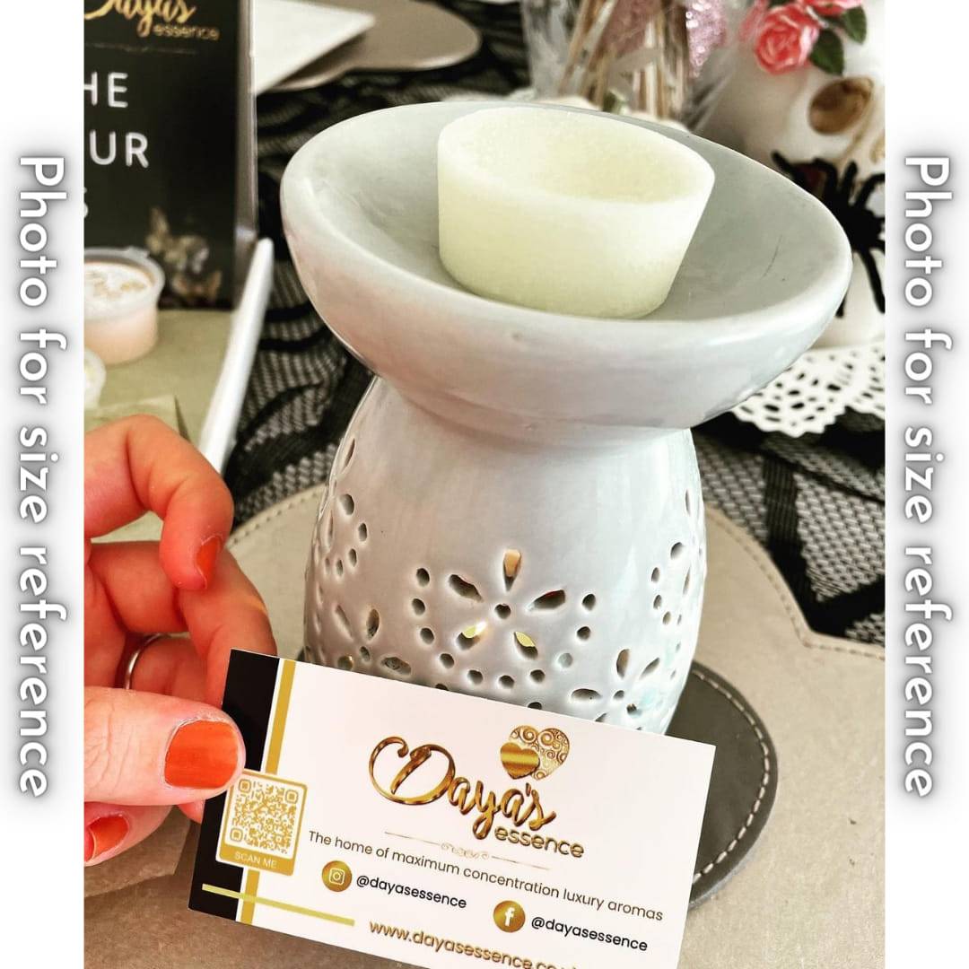 A light grey ceramic wax melt burner with a wax melt on top. The burner sits on a table and there is a hand holding a Daya's Essence business card in the foreground. The business card features the company logo, social media handles, and website.