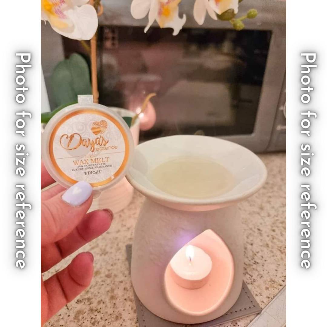 A hand holds a small white Daya's Essence wax melt labeled "FRESH" next to a ceramic wax melt warmer with a lit tealight candle. The wax melt is placed on top of the warmer.