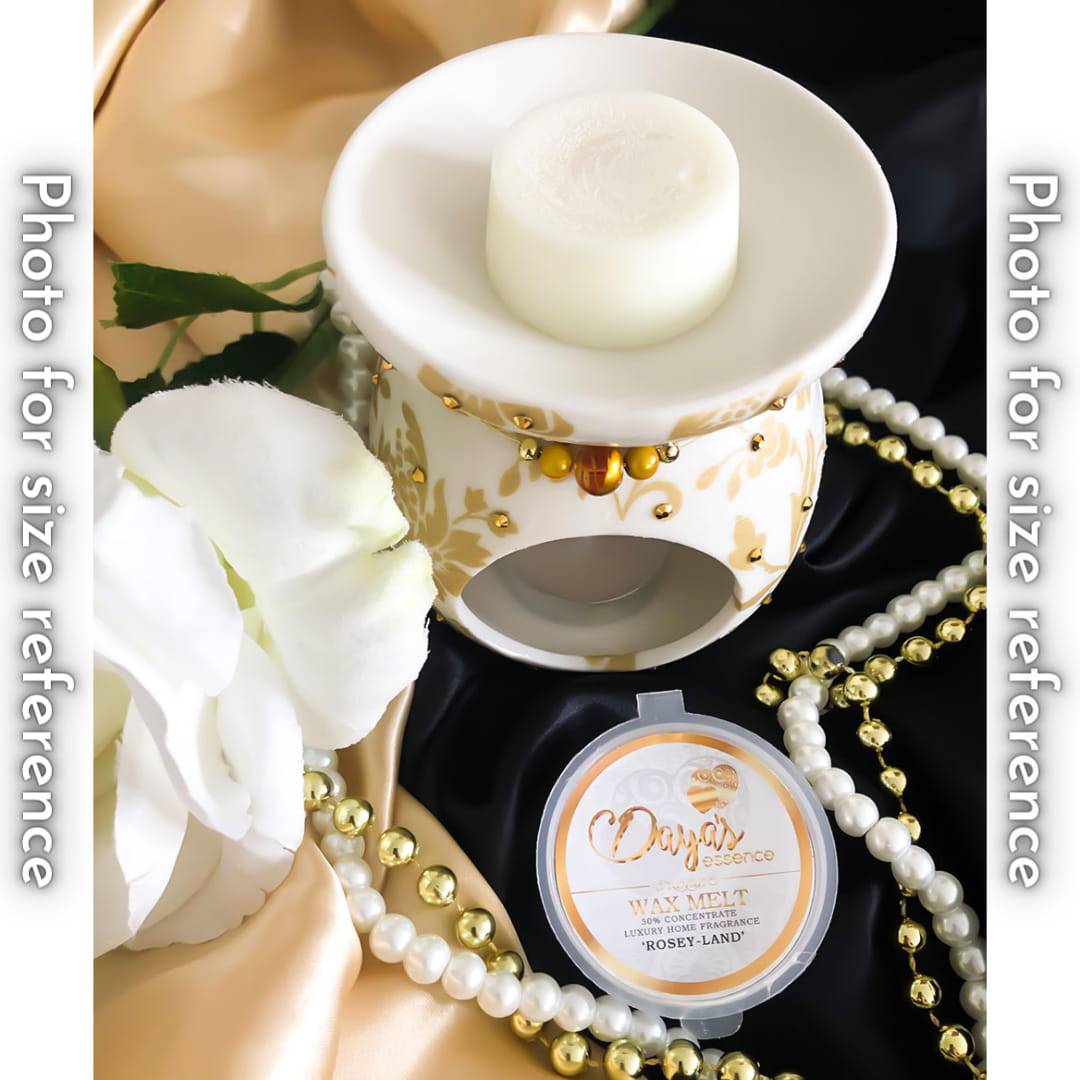A white ceramic wax melt warmer with a gold and white floral design holds a single white wax melt. The warmer is sitting on a silky fabric next to a string of pearls. A second wax melt, labeled 'Rosey-Land' is in its packaging in the foreground.