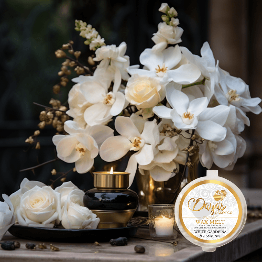 A "White Gardenia & Jasmine" wax melt by Dayas Essence sits on a reflective table next to a lit candle and a bouquet of white orchids. The warm glow of the candlelight illuminates the elegant floral arrangement, creating an ambiance of tranquility and luxury.