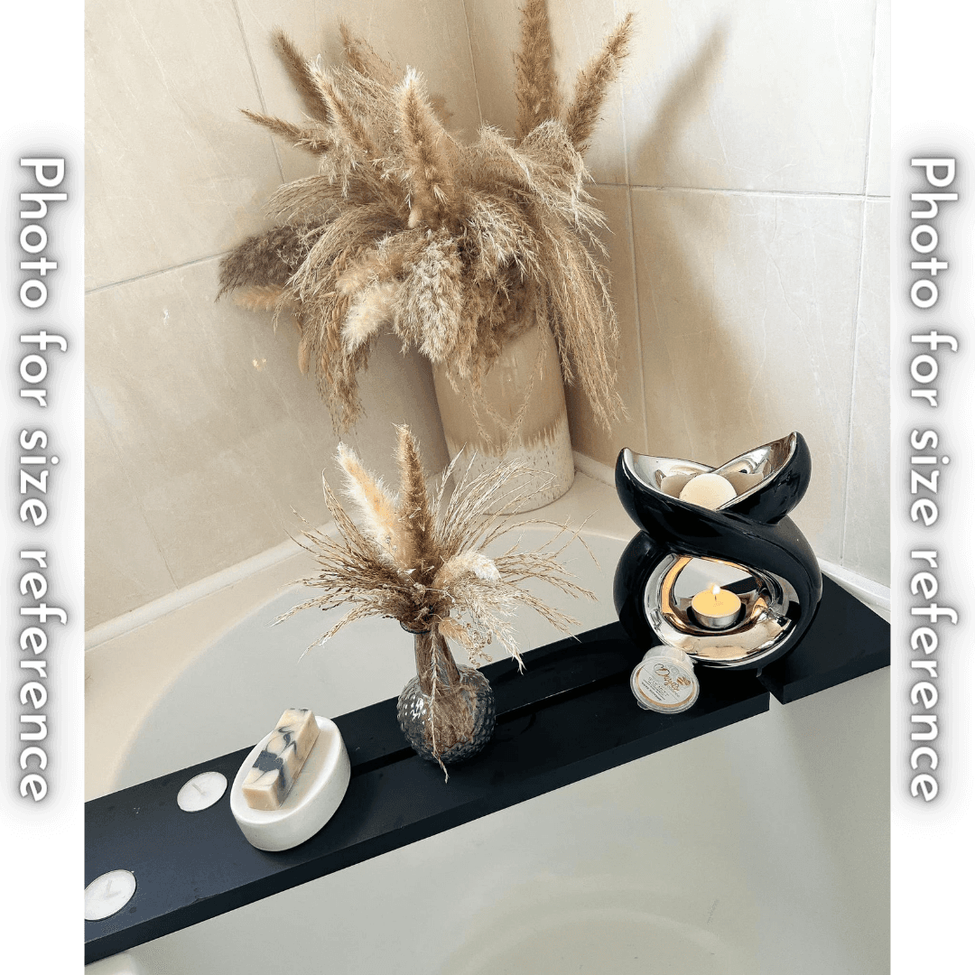 A black bath tray across a bathtub holds a bar of soap on a white dish, a vase with dried flowers, a black ceramic wax melt warmer with a lit candle, and a container of Daya's Essence wax melts.