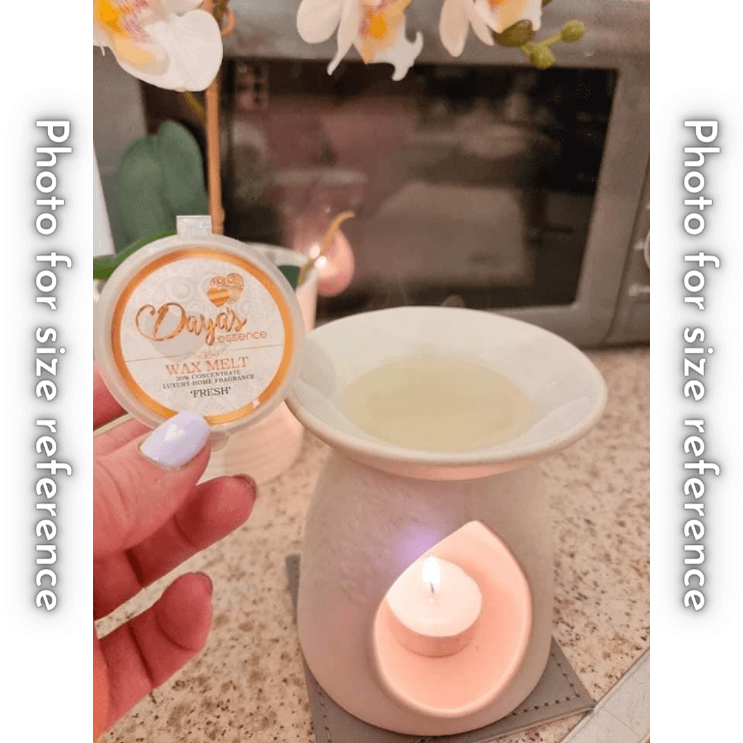 A hand holds a white wax melt labeled "Daya's Essence - FRESH" next to a white ceramic wax warmer with a lit tealight candle.