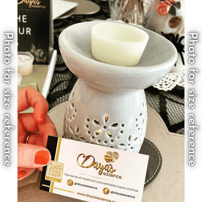 A light grey ceramic wax melt warmer with a white wax melt on top. The warmer sits on a surface, and a Daya's Essence business card is held in the foreground. The business card features the company logo, social media handles, and website.