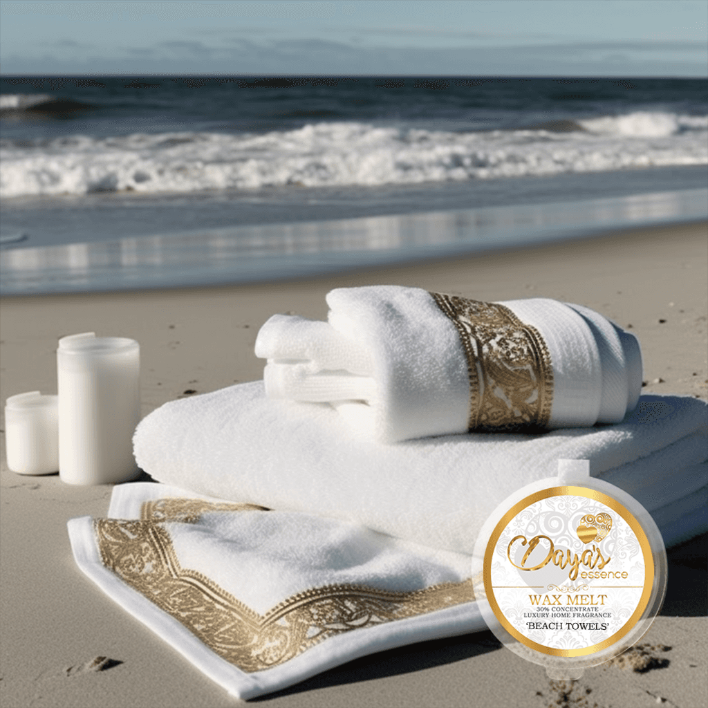 A white luxury hand towel with a gold leaf pattern border lies on a sandy beach with the ocean in the background. Next to it is a wax melt by Daya's Essence called 'Beach Towels'. There are two white candles next to the towel.