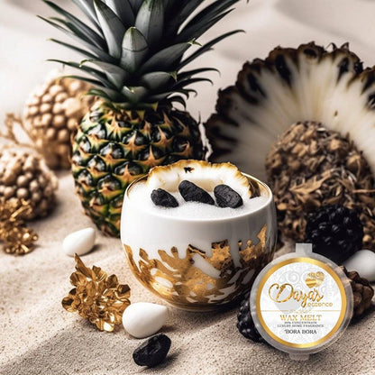 A white and gold container with a wax melt labeled "Bora Bora" by Daya's Essence sits on a white sand-like surface. The wax melt is surrounded by pineapple, black stones, and white pebbles, creating a tropical aesthetic.