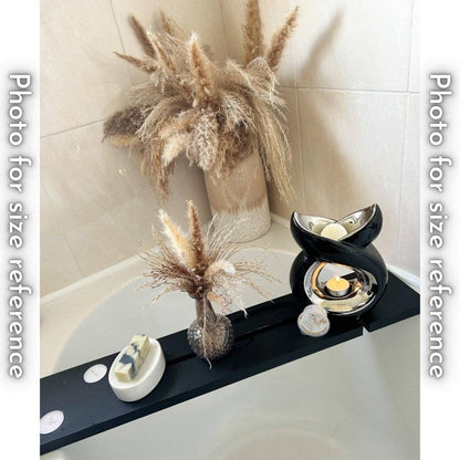 A black bath tray across a bathtub holds a bar of soap on a white dish, a vase with dried flowers, a black ceramic wax melt warmer with a lit candle, and a container of Daya's Essence wax melts.