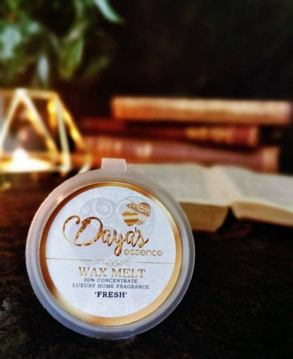 A single Daya's Essence wax melt labeled "Fresh" rests in a clear plastic clamshell container atop a stack of books, with a warm light glowing in the background, hinting at a cozy and fragrant atmosphere.