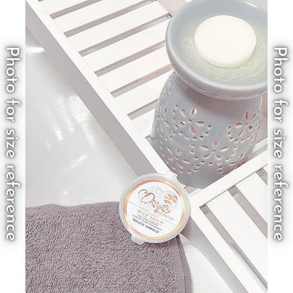 A light grey ceramic wax melt warmer with a white wax melt on top. The warmer sits on a white wooden tray, and a Daya's Essence "Beach Towels" wax melt is placed on a grey washcloth in the foreground.