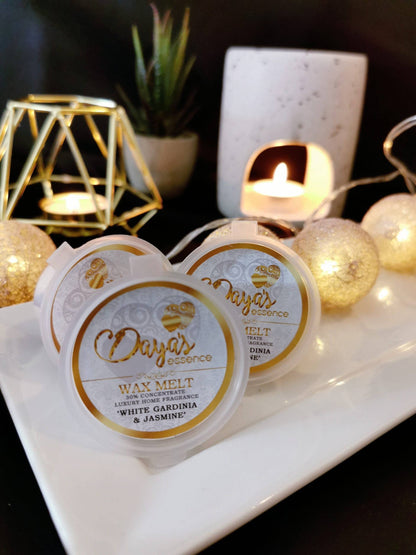 Three white containers labeled "Daya's Essence Wax Melt 30% Concentrate Luxury Home Fragrance 'White Gardenia & Jasmine'" are arranged on a white rectangular dish. The dish is surrounded by warm, glowing fairy lights and a tealight candle in a white ceramic holder. A geometric gold candle holder and a potted plant are blurred in the background.