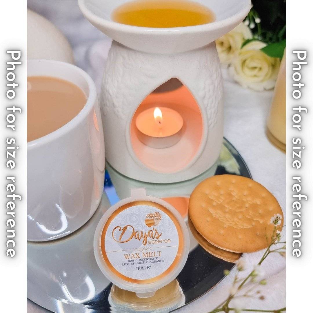 A white ceramic wax melt warmer with a tea light candle and melted wax sits next to a cup of coffee and a Daya's Essence wax melt labeled "Fate." A round cracker is placed in front to show the size of the wax melt.